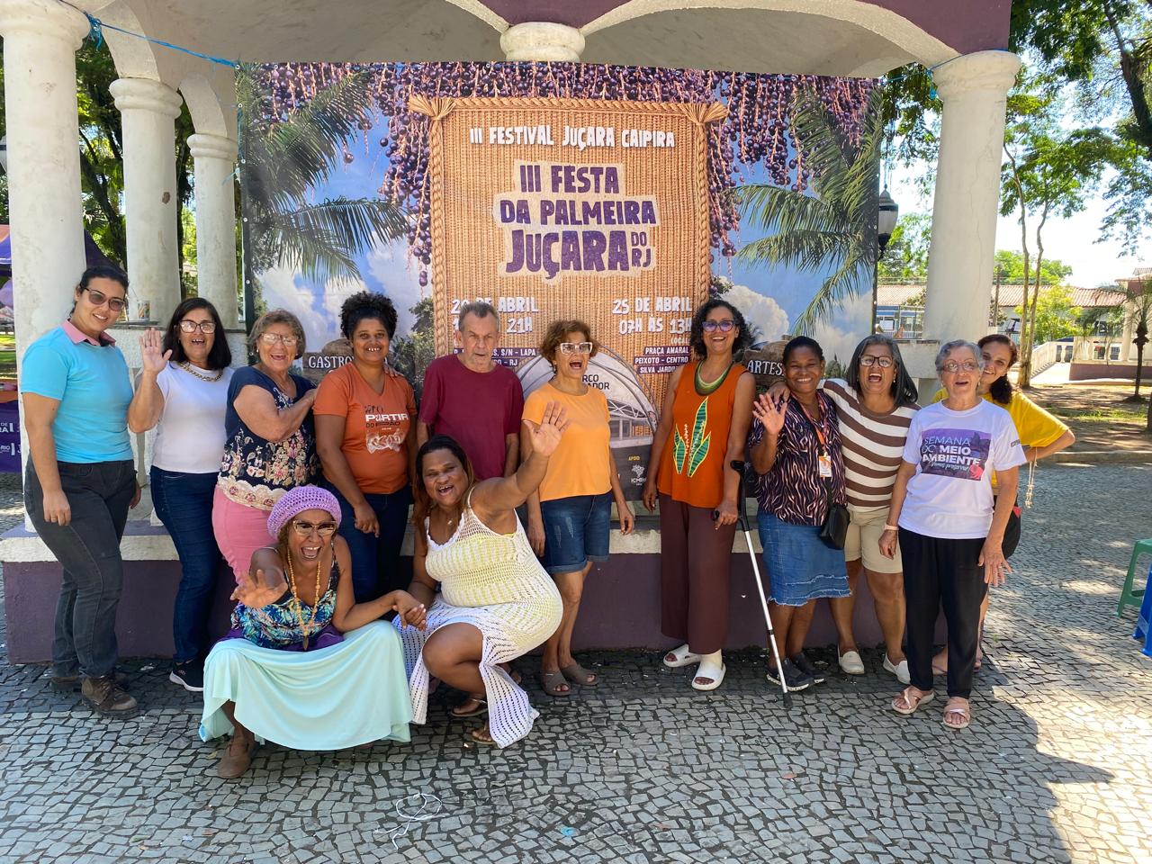Grupo de participantes posando em frente ao cartaz da III Festa da Palmeira Juçara na Praça Amaral Peixoto