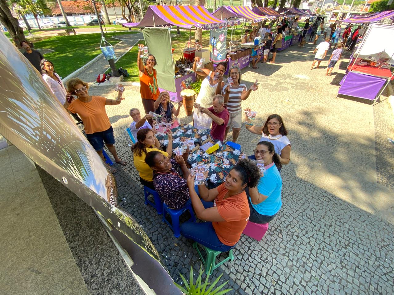 Vista aérea da roda de artesãs montando colares na feira em Silva Jardim, com barracas coloridas ao fundo