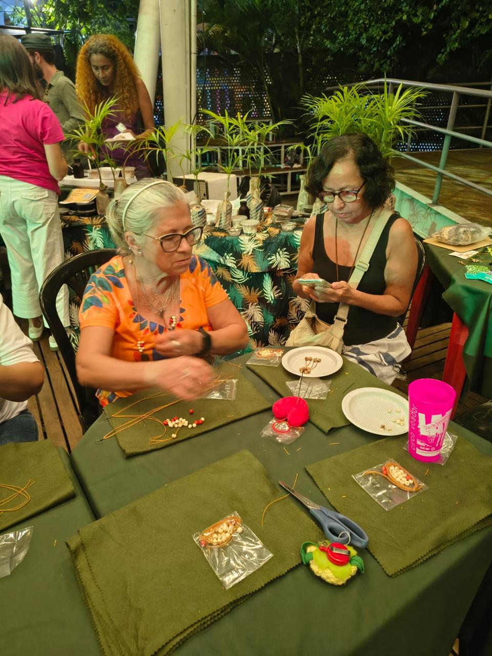 Mulheres confeccionando peças com sementes de juçara cercadas por mudas da palmeira